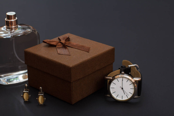 Κολωνία για άνδρες, καφέ συσκευασία δώρου, μανικετόκουμπα και ρολόι με ένα μαύρο δερμάτινο λουράκι σε μαύρο φόντο. ΑΞΕΣΟΥΑΡ ΑΝΔΡΙΚΑ. - Φωτογραφία, εικόνα