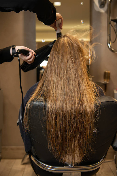 Перукар сушить красиве довге коричневе волосся з феном і щіткою для волосся, поки дівчина сидить у кріслі в салоні краси. Створення стилю
. - Фото, зображення