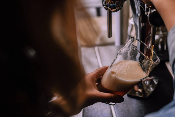 Κορίτσι ρίχνει μια πίντα του μαύρη μπύρα σε ένα μπαρ μπύρα σκάφη. Τοπική παμπ. Λαμπερά beer βρύσες στο παρασκήνιο. Φωτεινό ηλιόλουστο απόγευμα. Φαίνονται από τον ένα ώμο - Φωτογραφία, εικόνα