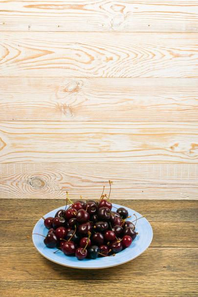 Round blue plate of fresh ripe cherries on old wooden background for design montage. Vintage wood table desk board with overripe cherries or sweet berries pile close up with copy space - Фото, изображение