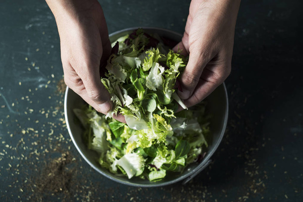 Nahaufnahme eines jungen kaukasischen Mannes, der gerade dabei ist, einen Salat mit einer Mischung aus verschiedenen Salatblättern wie Römersalat, Endivien oder Rucola in einer rustikalen Metallschale zuzubereiten. - Foto, Bild