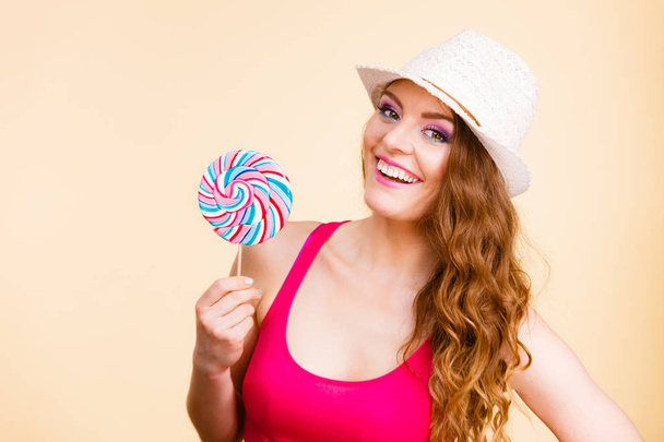 Beautiful woman wearing red tshirt summer hat holding big lollipop candy in hand. Sweet food fun concept. Studio shot on bright beige - Foto, afbeelding