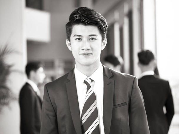 Porträt eines jungen asiatischen Geschäftsmannes, der auf die Kamera zeigt und sie lächelnd ansieht, Kollegen, die im Hintergrund chatten, schwarz-weiß. - Foto, Bild