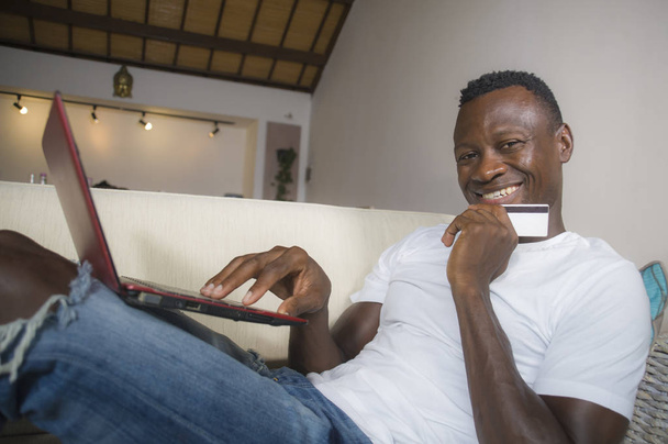 lifestyle portrait of young happy and attractive black afro American man using credit card and laptop computer relaxed and cheerful at living room sofa couch internet banking and online shopping - 写真・画像