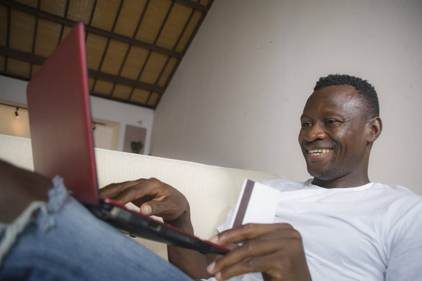 lifestyle portrait of young happy and attractive black afro American man using credit card and laptop computer relaxed and cheerful at living room sofa couch internet banking and online shopping - Zdjęcie, obraz