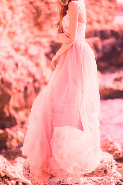 beautiful girl stands sideways in a pink dress of tulle on the nature of the sea with rocks. The concept of fashion colors 2019, living coral - Photo, Image