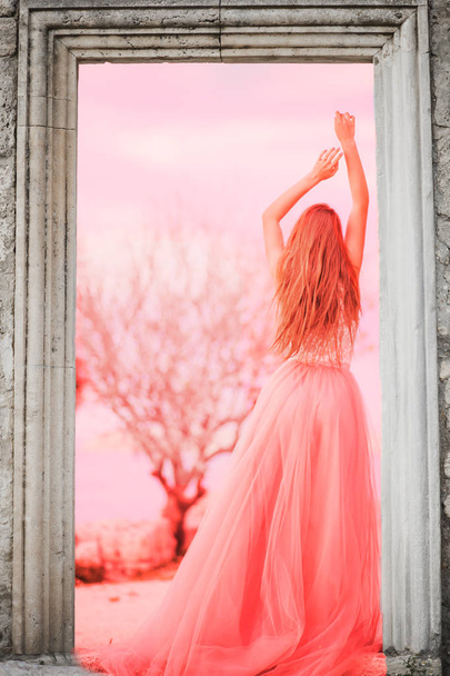 Woman in pink long dress at the sea is back at the old castle. The concept of fashion colors 2019, living coral - Photo, image