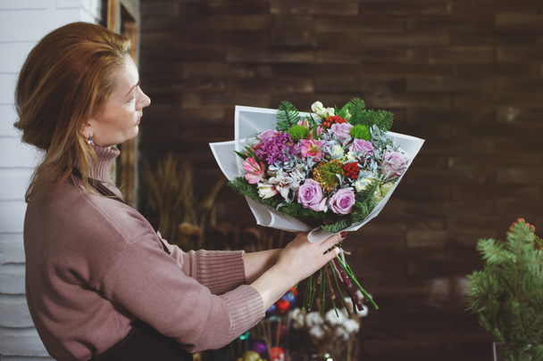 Ανθοπωλείο αρκετά μεσήλικας γυναίκα στην ποδιά κάνει μια ανθοδέσμη στο κομμωτήριο λουλούδι για ένα γιορτινό δώρο για ένα γάμο ή την επέτειό σας. - Φωτογραφία, εικόνα