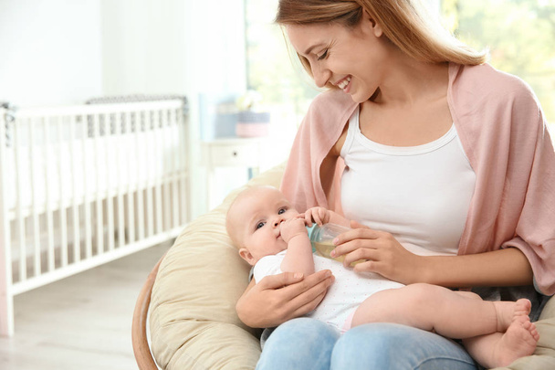 Lovely mother giving her baby drink from bottle in room. Space for text - 写真・画像