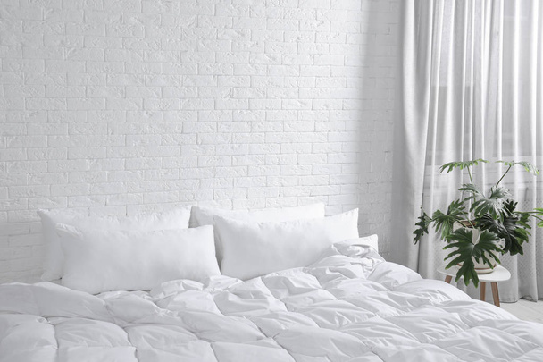 Large comfortable bed with pillows and blanket near white brick wall indoors. Stylish interior - Foto, afbeelding