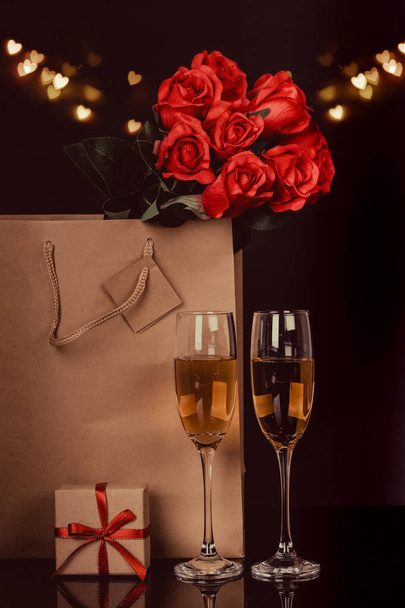 Champagne bottle in a paper bag, two wine glasses and  red roses on black background with lights in the background. Love, Valentines day concept - Foto, imagen