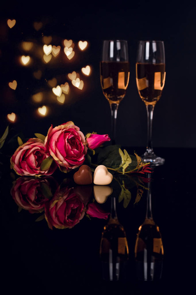 Two glasses of champagne, roses and two heart shape chocolates on black background with lights in the background. Love, Valentines day concept - Photo, image