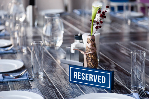 Крупный план деревянная бело-голубая прямоугольная пластина со словом Зарезервировано стоя на сером винтажном столе в ресторане рядом с установкой, салфетки, соль перец, ваза с цветами, очки, керамические белые тарелки
 - Фото, изображение