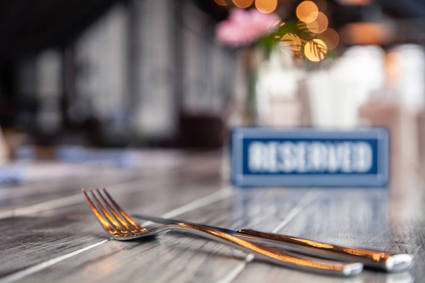 Closeup metal fork and knife near wooden blue white rectangular plate with the word Reserved standing on gray vintage table in restaurant. Soft focus, blurred object. Concept closed banquet - Photo, Image