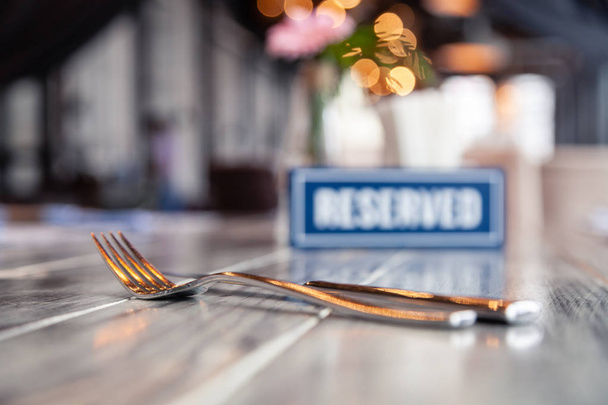 Closeup metal fork and knife near wooden blue white rectangular plate with the word Reserved standing on gray vintage table in restaurant. Soft focus, blurred object. Concept closed banquet - Photo, Image
