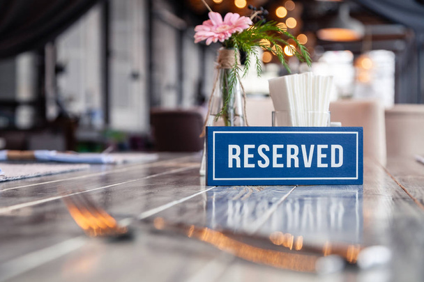 Closeup de madeira azul placa retangular branca com a palavra Reservado de pé sobre mesa vintage cinza no restaurante perto de configuração, guardanapos, garfo, faca, vaso com flores, óculos, placas brancas de cerâmica
 - Foto, Imagem