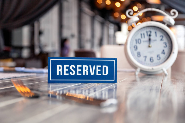 Closeup wooden blue white rectangular plate with the word Reserved standing on gray vintage table in restaurant near to setting, napkins, fork, knife, clock - Photo, Image