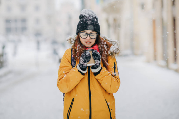 Girl in yellow coat drinking coffee outdoors. Lonely woman stands on a snowy winter street in the city - Photo, Image