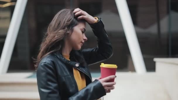 Beautiful young woman with an attractive appearance, adorable look and autumn jacket in the city. Amazing view of business lady with a cup of coffee walking down the city street - Πλάνα, βίντεο