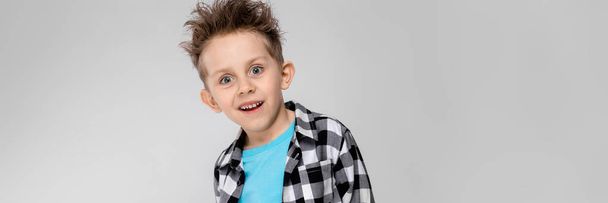 A handsome boy in a plaid shirt, blue shirt and jeans stands on a gray background. The boy folded his arms behind his back - Foto, imagen