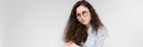 Portrait of a young girl in glasses. Beautiful young girl on a gray background. The girl is holding glasses in her hands. The girl folded her arms. - Photo, Image