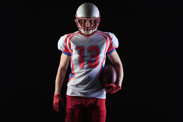 American football player wearing helmet posing with ball on black background - Photo, Image