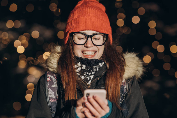 Beautiful caucasian woman using smart phone hand hold outdoor in the city night, smiling, face illuminated screenlight - Fotó, kép