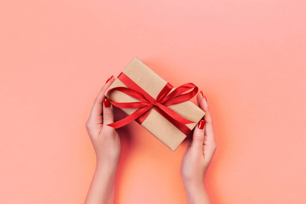 Woman hands give wrapped valentine or other holiday handmade present in paper with red ribbon. Present box, decoration of gift on Coral color table, top view with copy space. - Φωτογραφία, εικόνα