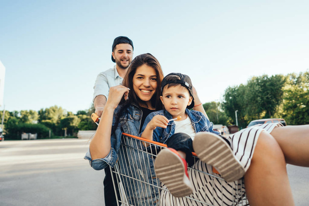 Young dad carries mom and son in a cart on the parking lot. The family is having fun. - 写真・画像