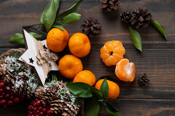 The atmosphere of Christmas and New Year: Fresh tangerine with green leaves on a dark wooden background. Mandarin peeled and disassembled into slices, cinnamon sticks, cones, tangerine peels, leaves. - Foto, afbeelding