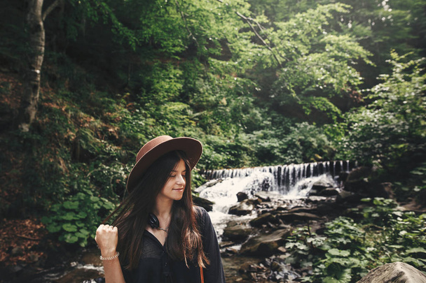 Stylish hipster girl in hat traveling in woods. Young woman with beautiful hair exploring in summer forest, looking at river. Travel and wanderlust concept. Space for text - Foto, Imagem