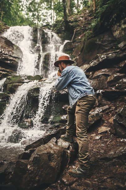 Stylish young man in hat traveling in mountains. Hipster man with camera taking photo of beautiful waterfall, exploring summer forest. Travel and wanderlust concept. Space for text. - Photo, Image