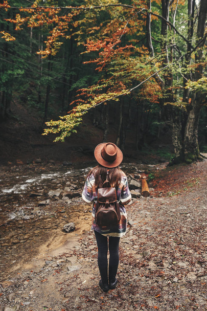 Stylish hipster girl in hat traveling in mountains. Young woman with backpack exploring and walking in summer forest. Travel and wanderlust concept. Space for text. Back view - Фото, изображение