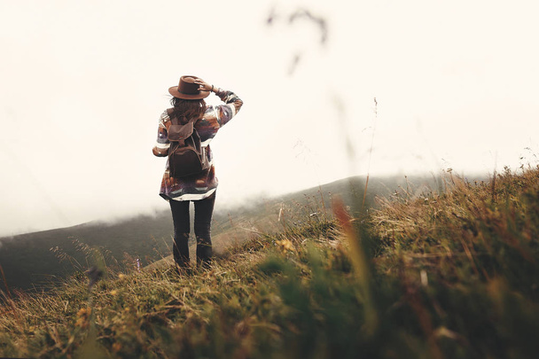 Stylish hipster girl in hat walking on top of mountains. Happy young woman with backpack exploring misty mountains. Travel and wanderlust concept. Amazing atmospheric moment - Fotó, kép