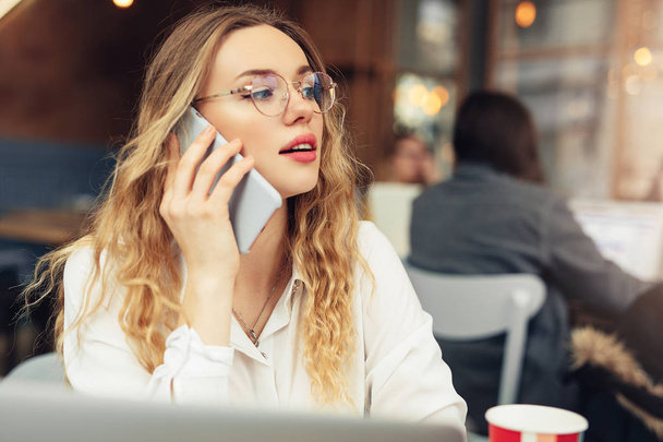 Woman Wear Glasses, Talk At Phone, Drink Her Hot Coffee While Work In Cafe On Her Laptop. Portrait Of Stylish Smiling Woman In Winter Clothes Drinking Hot Coffee And Work At Laptop. Female Winter Style. - Image - Foto, imagen