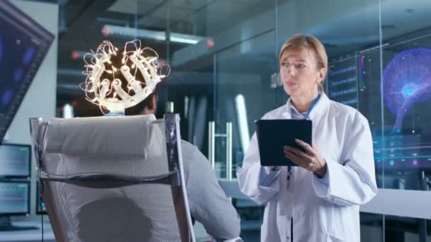 Człowiek sobie Brainwave skanowania zestawu słuchawkowego siedzi na krześle, podczas gdy naukowiec z Tablet komputer nadzoruje proces. W mózgu nowoczesne badania laboratoryjne monitory Pokaż Eeg czytania i Model mózgu. - Materiał filmowy, wideo