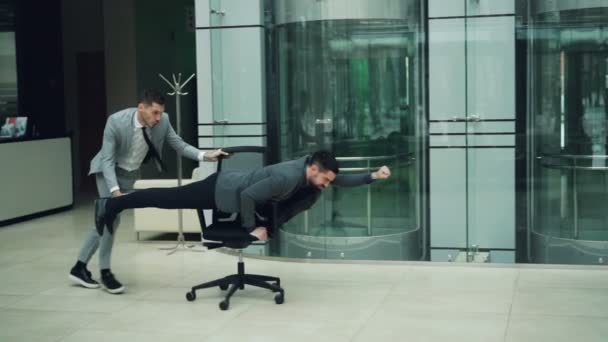 Playful business people are having fun in office lobby riding chair and playing racing game on rolling armchair. Emotional youth, joyful adults and work concept. - Footage, Video