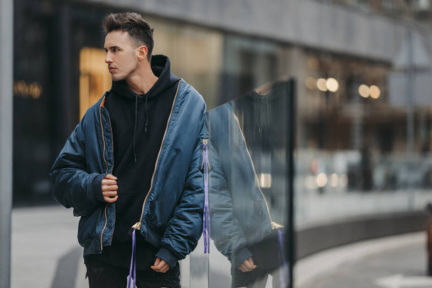 Fashionable man walk on the street near buildings. Wear blue jacket and all black. Winter, autumn outfit. Jacket with blouse and black sneakers. - Fotó, kép