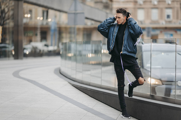 ファッショナブルな男は、建物の近くの通りを歩きます。青いジャケットとすべての黒を着用します。冬、秋服。ブラウスと黒のスニーカーとジャケット. - 写真・画像