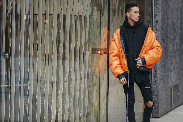 Fashionable man stand on the street near buildings. Wear orange jacket and all black. Winter, autumn outfit. Jacket with blouse and black sneakers. - Foto, Imagem
