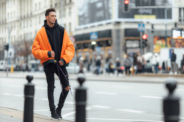 おしゃれな男は、建物近くの路上に立っています。オレンジ色のジャケットとすべての黒を着用します。冬、秋服。ブラウスと黒のスニーカーとジャケット. - 写真・画像