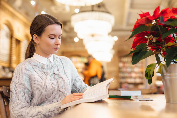 Clever girl sit at cafe and read a interesting book with boring face. Wear white sweater. Winter, autumn outfit. - 写真・画像