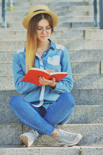 Mladá studentka v klobouku a džínové košili píše nápady do sešitu, sedí na schodech na ulici a usmívá se do kamery. - Fotografie, Obrázek
