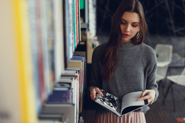 Young fashion woman hold her interesting book, stand near the wall of books and read. Look around and smile to camera. Copy space. - Image - Zdjęcie, obraz