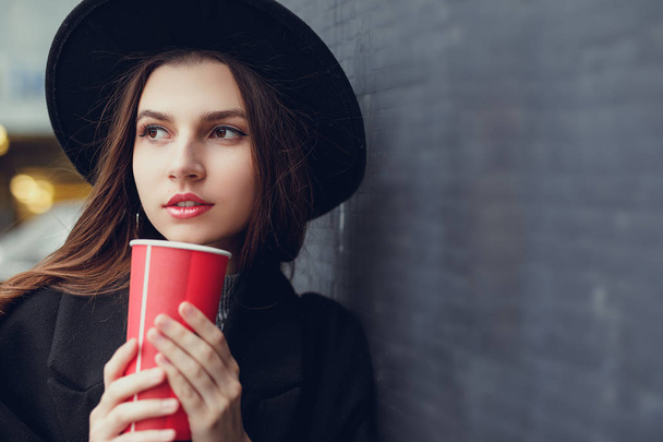 Young fashion woman hold her hot cup of coffee, stand near the wall wear black hat look around and smile to camera. Copy space. - Image - Foto, Bild