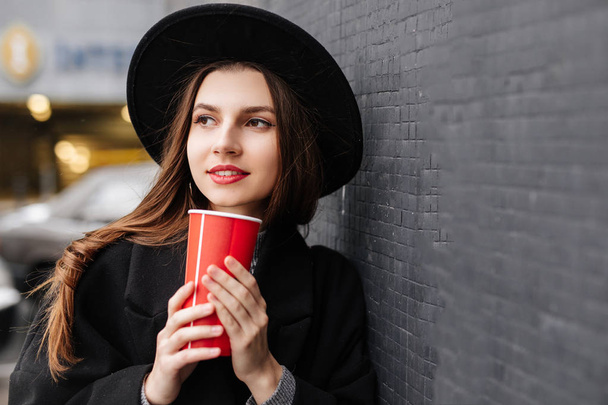 Young fashion woman hold her hot cup of coffee, stand near the wall wear black hat look around and smile to camera. Copy space. - Image - Valokuva, kuva