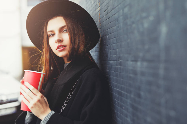 Jeune femme de mode tenir sa tasse de café chaud, se tenir près du mur porter chapeau noir regarder autour et sourire à la caméra. Espace de copie. - Image
 - Photo, image