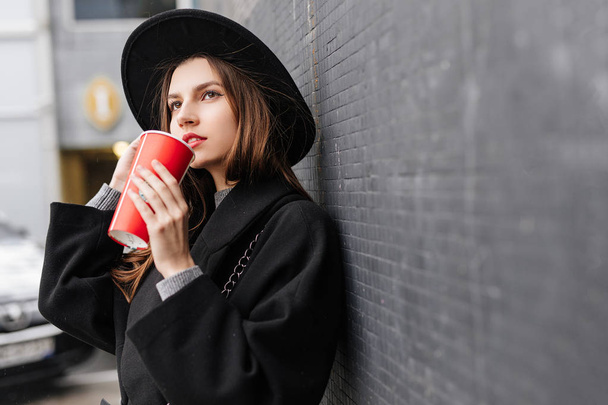 Young fashion woman hold her hot cup of coffee, stand near the wall wear black hat look around and smile to camera. Copy space. - Image - Photo, Image