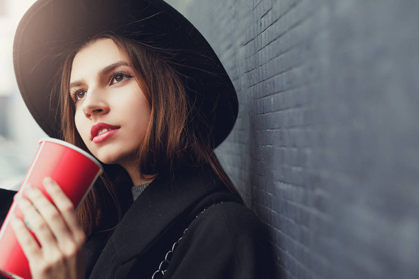 Young fashion woman hold her hot cup of coffee, stand near the wall wear black hat look around and smile to camera. Copy space. - Image - Fotografie, Obrázek