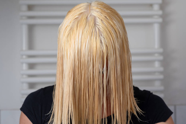 Блондинка с мокрыми волосами длиной плеч наклоняется вперед после мытья с шампунем в ванной комнате с видом на ее волосы
 - Фото, изображение
